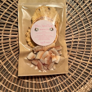 May include: A transparent bag containing dried pineapple and orange slices, along with coconut flakes. The bag has a brown paper backing and a label that reads "Mom's Tropical Escape Simmer Pot". It rests on a woven wicker placemat.