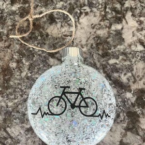 May include: Clear glass Christmas ornament filled with iridescent glitter. A black bicycle graphic and a heartbeat line are featured on the front. The ornament has a silver metal cap and a twine loop for hanging.