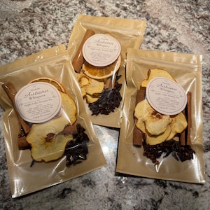 May include: Three clear plastic pouches containing dried apple slices, cinnamon sticks, and whole cloves. Each pouch features a round white label with the text Autumn Whispers Apple Cider Simmer Pot and instructions for use on the front.