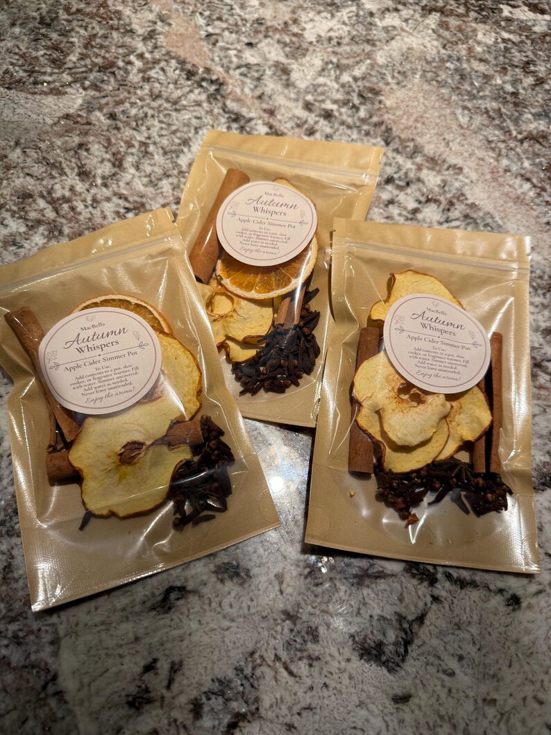 May include: Three clear plastic pouches containing dried apple slices, cinnamon sticks, and whole cloves. Each pouch features a round white label with the text Autumn Whispers Apple Cider Simmer Pot and instructions for use on the front.