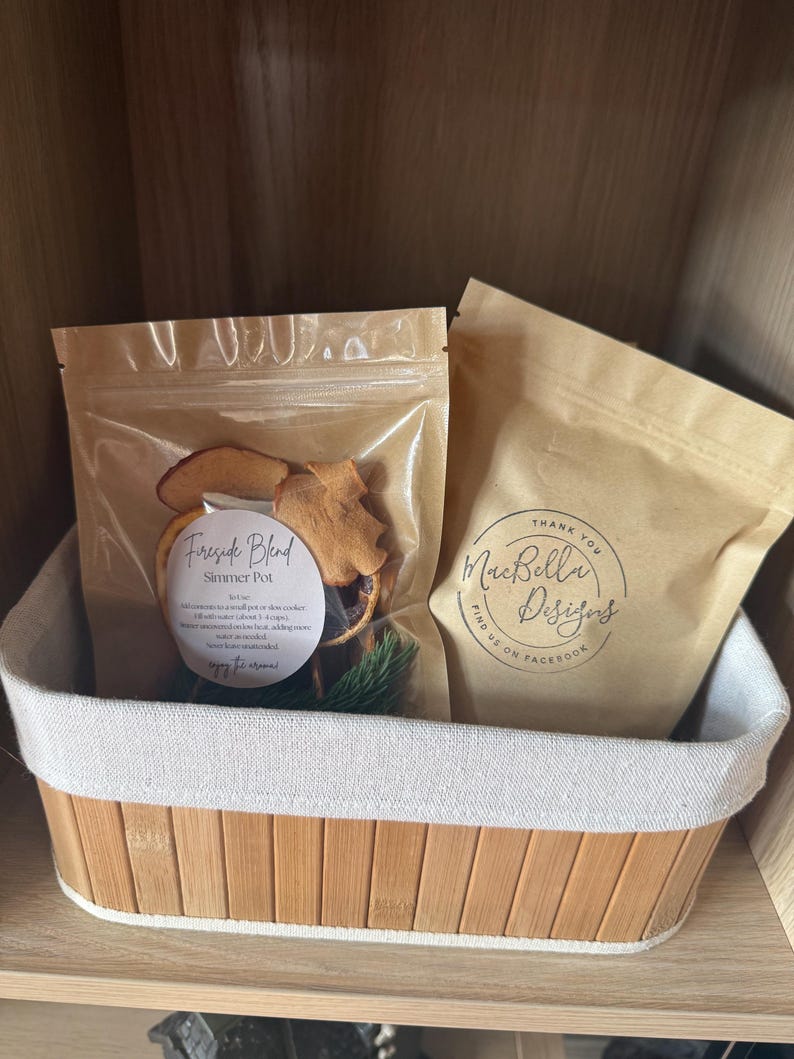 May include: Two brown paper pouches in a bamboo basket. The left pouch is clear-fronted, containing dried fruit and pine, labeled 'Fireside Blend Simmer Pot'. The right pouch is opaque brown paper with the text 'Thank You MacBella Designs'.