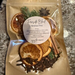 Peut inclure: Un sachet transparent rempli d'un kit de pot-pourri "Fireside Blend". Le kit comprend des tranches d'orange séchées, de la badiane, des bâtons de cannelle, des clous de girofle et des brins de pin. Le texte sur l'étiquette dit "enjoy the aroma!"