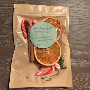 May include: A clear plastic pouch with a brown paper backing containing a Christmas Spirit Simmer Pot kit. The kit includes dried orange slices, cinnamon sticks, small candy canes, and evergreen sprigs, with a label providing instructions.