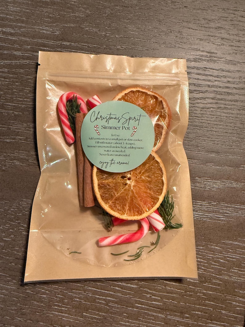 May include: A clear plastic pouch with a brown paper backing containing a Christmas Spirit Simmer Pot kit. The kit includes dried orange slices, cinnamon sticks, small candy canes, and evergreen sprigs, with a label providing instructions.
