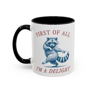 May include: A white ceramic mug with a black handle and interior. The mug features a blue illustration of a raccoon in a dancing pose, centered between the red text "FIRST OF ALL" at the top and "I'M A DELIGHT" at the bottom of the design.