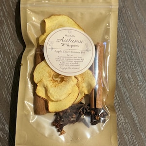 May include: A clear plastic pouch containing dried apple slices, two cinnamon sticks, star anise, and cloves. A circular label on the front reads MacBella Autumn Whispers Apple Cider Simmer Pot, with instructions to add contents to a pot, slow cooker, or fragrance warmer.