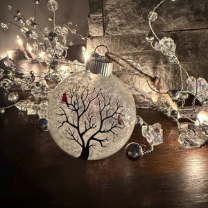 May include: A clear glass Christmas ornament with a black tree design and two birds. The ornament is decorated with silver and clear beads and lights, creating a festive holiday scene.