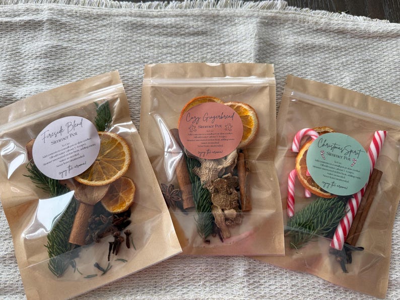 May include: Three clear pouches containing dried simmer pot ingredients. The left pouch labeled Fireside Blend features orange slices, cinnamon sticks, and pine needles. The center pouch labeled Cozy Gingerbread includes ginger, cinnamon, and pine. The right pouch labeled Christmas Spirit contains orange slices, pine, cinnamon, and candy canes.