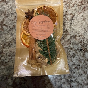 May include: A clear plastic pouch containing a Cozy Gingerbread Simmer Pot mix, featuring dried orange slices, a star anise, cinnamon sticks, a pine sprig, and dried ginger pieces. A round label on the front displays the text Cozy Gingerbread Simmer Pot.