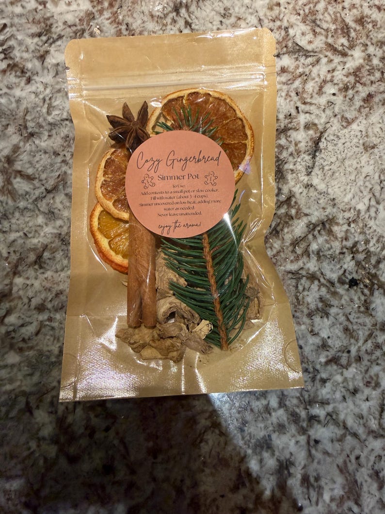 May include: A clear plastic pouch containing a Cozy Gingerbread Simmer Pot mix, featuring dried orange slices, a star anise, cinnamon sticks, a pine sprig, and dried ginger pieces. A round label on the front displays the text Cozy Gingerbread Simmer Pot.