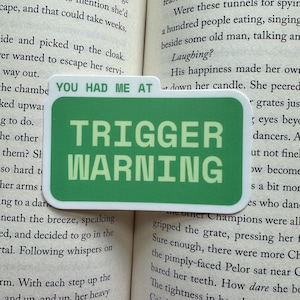 May include: A green sticker with the text "YOU HAD ME AT TRIGGER WARNING" in a bold, sans-serif font. The sticker is rectangular with rounded corners and is placed on an open book.