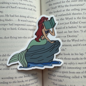 May include: A die-cut sticker depicting a cartoon mermaid reading a book. The mermaid has red hair, a seashell top, and a green and blue tail, sitting on a rock. The sticker is placed on an open book.