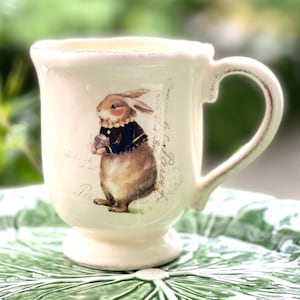 May include: A white ceramic mug with a handle features a hand-painted illustration of a rabbit wearing a blue jacket and holding a small gift. The mug has a distressed finish and the text "Paris" and "Alice" is written on the side.