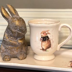 May include: A white ceramic mug with a brown bunny illustration and the text "So Very Pretty" on the side. The mug is on a white tray with a gold bunny figurine.