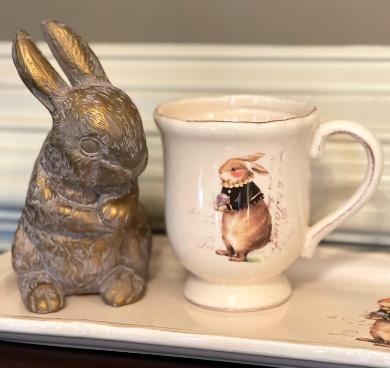 May include: A white ceramic mug with a brown bunny illustration and the text "So Very Pretty" on the side. The mug is on a white tray with a gold bunny figurine.