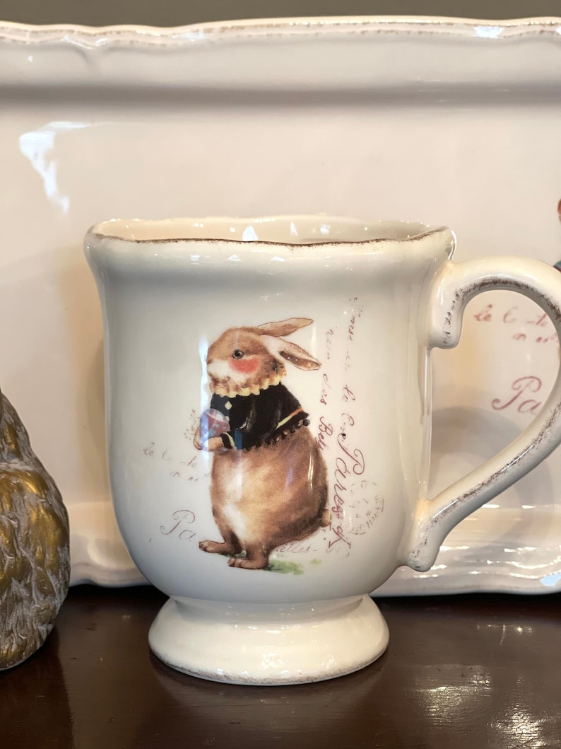 May include: White ceramic mug featuring a brown rabbit illustration and the text "By Dress of the Day" in pink. The mug has a handle and a rounded base.