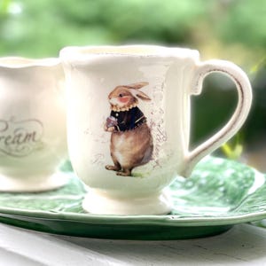 May include: A white ceramic teacup with a handle features a brown rabbit illustration wearing a black jacket and holding a flower. The text "Tea" and "La Boutique" are visible on the cup.
