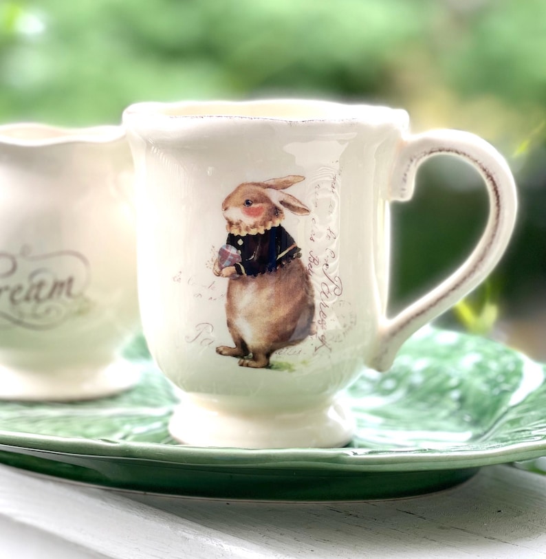 May include: A white ceramic teacup with a handle features a brown rabbit illustration wearing a black jacket and holding a flower. The text "Tea" and "La Boutique" are visible on the cup.
