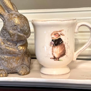 May include: A white ceramic mug with a brown bunny illustration and a handle. The mug is sitting on a white ceramic tray with a brown bunny illustration. A gold bunny figurine is on the left side of the tray.