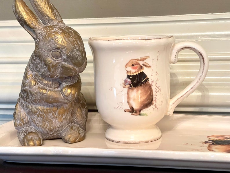 May include: A white ceramic mug with a brown bunny illustration and a handle. The mug is sitting on a white ceramic tray with a brown bunny illustration. A gold bunny figurine is on the left side of the tray.