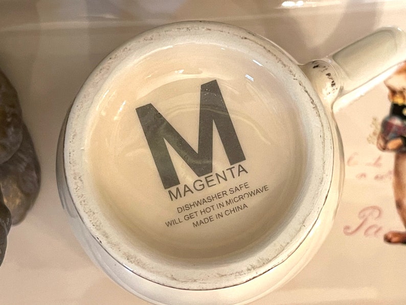 May include: White ceramic mug with a black letter "M" and the word "MAGENTA" printed on the side. The mug also has the text "Dishwasher Safe, Will Get Hot in Microwave, Made in China".