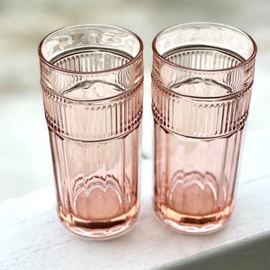 May include: Two pink glass tumblers with a ribbed design. The glasses are empty and sitting on a white surface.
