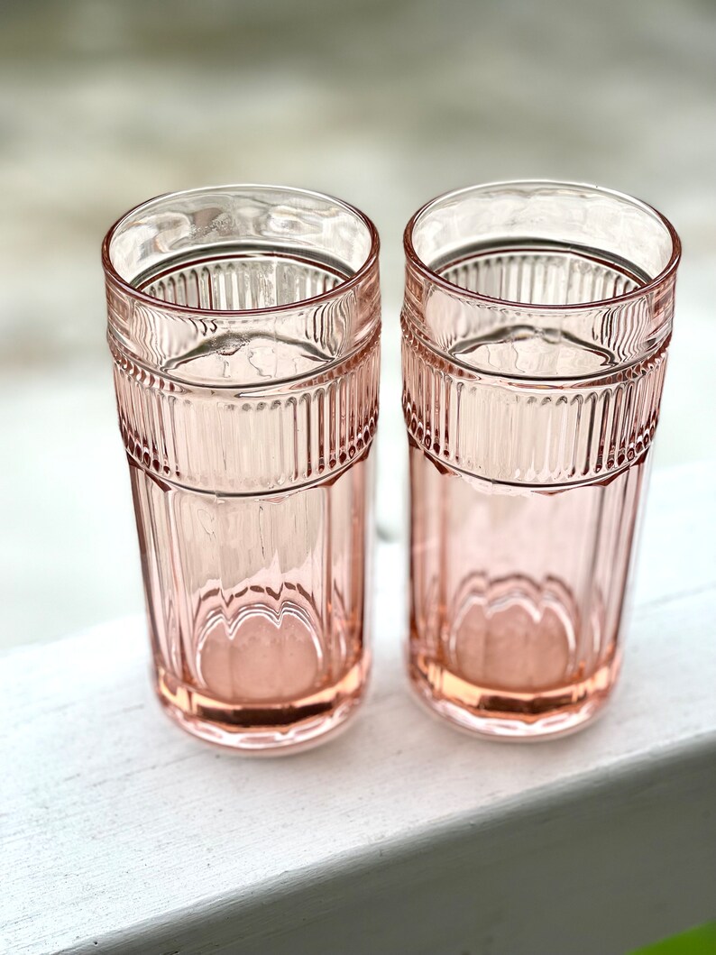 May include: Two pink glass tumblers with a ribbed design. The glasses are empty and sitting on a white surface.