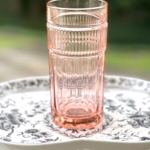 May include: A pink glass drinking glass with a ribbed design. The glass is sitting on a white tray with a black floral pattern.