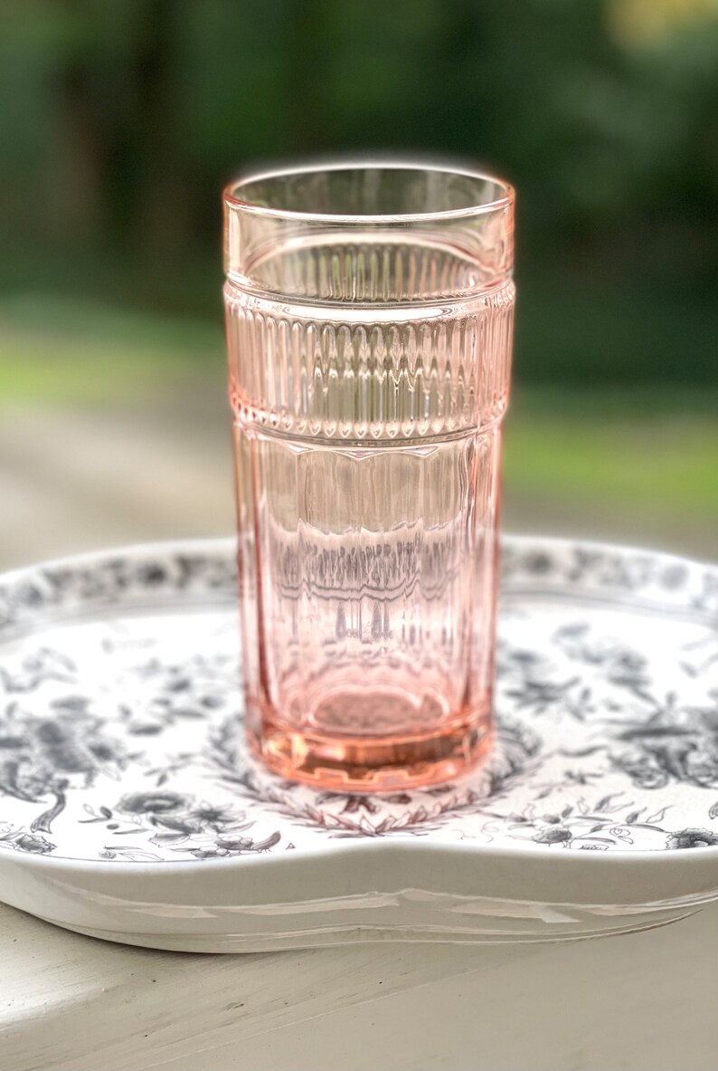 May include: A pink glass drinking glass with a ribbed design. The glass is sitting on a white tray with a black floral pattern.
