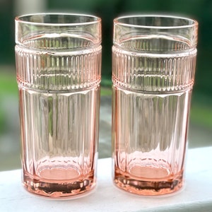 May include: Two clear pink glass tumblers with a ribbed design. The glasses are empty and sitting on a white surface.