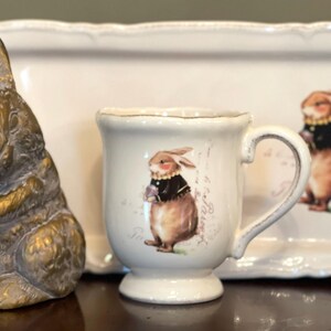 May include: A white ceramic mug with a handle features a brown rabbit wearing a black waistcoat and holding a flower. The mug is sitting on a white ceramic tray with a brown rabbit wearing a black waistcoat and holding a flower. The tray has a scalloped edge.