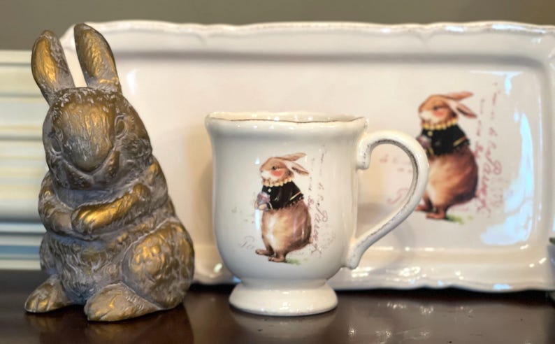 May include: A white ceramic mug with a handle features a brown rabbit wearing a black waistcoat and holding a flower. The mug is sitting on a white ceramic tray with a brown rabbit wearing a black waistcoat and holding a flower. The tray has a scalloped edge.