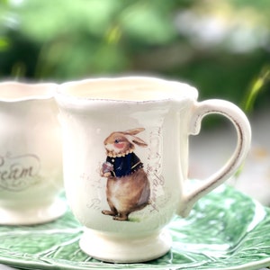 May include: A white ceramic mug with a handle features a brown rabbit wearing a blue jacket and holding a flower. The mug has a distressed look and is decorated with a floral pattern and text in French.