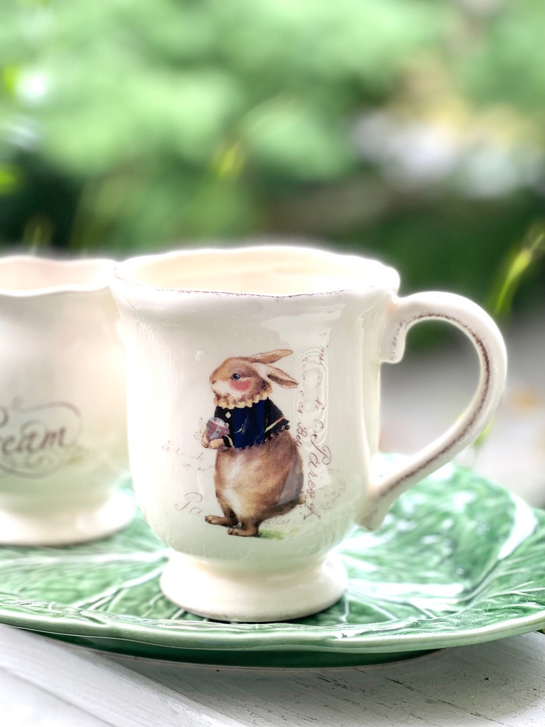 May include: A white ceramic mug with a handle features a brown rabbit wearing a blue jacket and holding a flower. The mug has a distressed look and is decorated with a floral pattern and text in French.