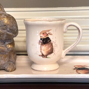 May include: A white ceramic mug featuring a brown bunny wearing a black jacket and holding a flower. The mug has a handle and a decorative design with the text "Paris" on the front.
