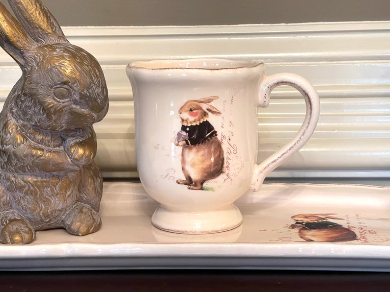 May include: A white ceramic mug featuring a brown bunny wearing a black jacket and holding a flower. The mug has a handle and a decorative design with the text "Paris" on the front.