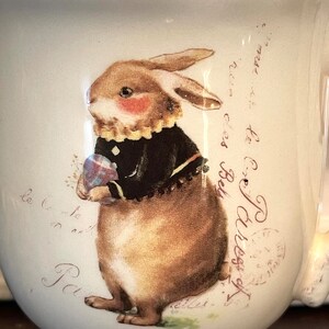 May include: White ceramic mug featuring a brown bunny holding a blue and pink egg. The bunny is wearing a black jacket and a white collar. The mug has a scalloped rim and the text "rue des Bely to Ex Paress of le to le Pa" is printed on the side.