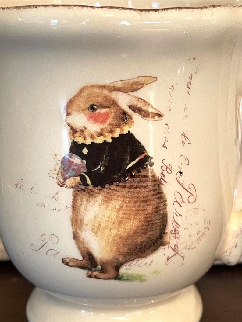May include: White ceramic mug featuring a brown bunny holding a blue and pink egg. The bunny is wearing a black jacket and a white collar. The mug has a scalloped rim and the text "rue des Bely to Ex Paress of le to le Pa" is printed on the side.