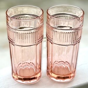May include: Two pink glass tumblers with a ribbed design. The glasses are empty and sitting on a white surface.