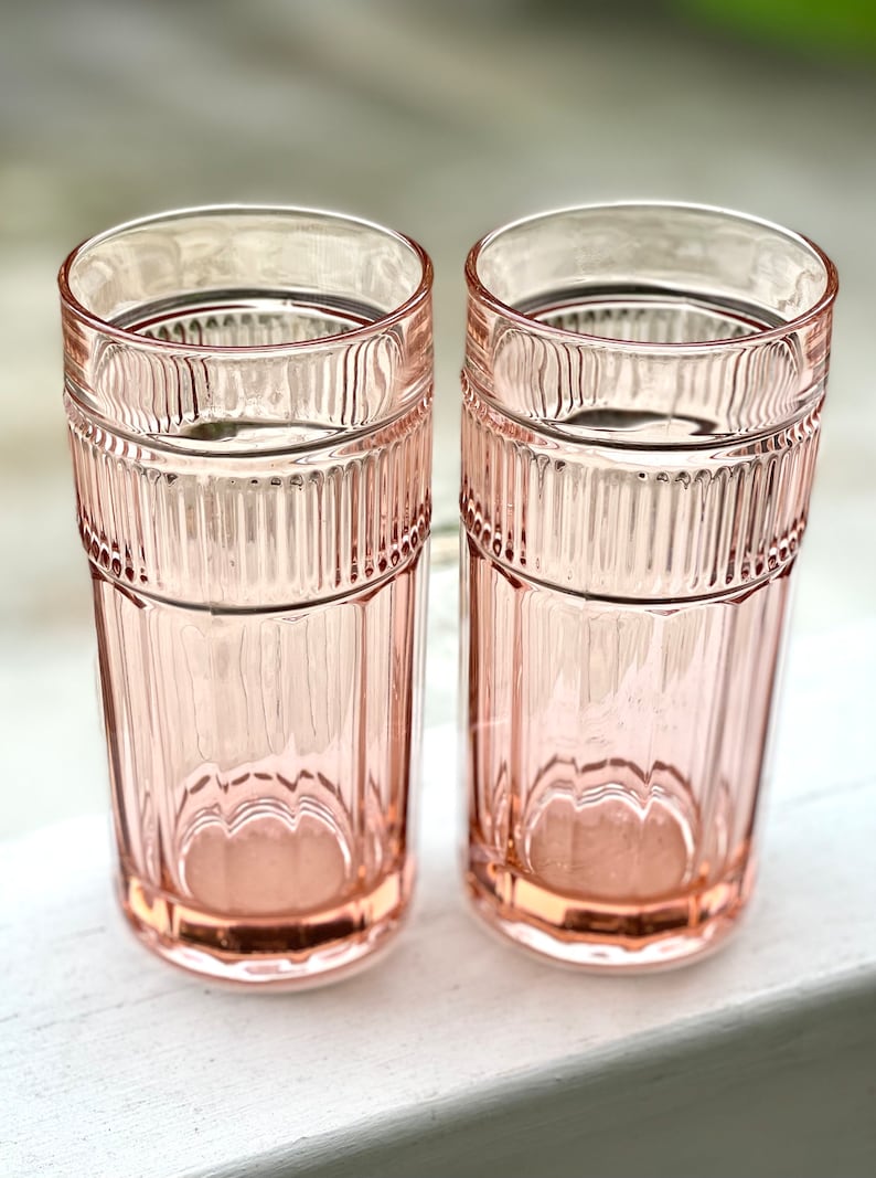 May include: Two pink glass tumblers with a ribbed design. The glasses are empty and sitting on a white surface.