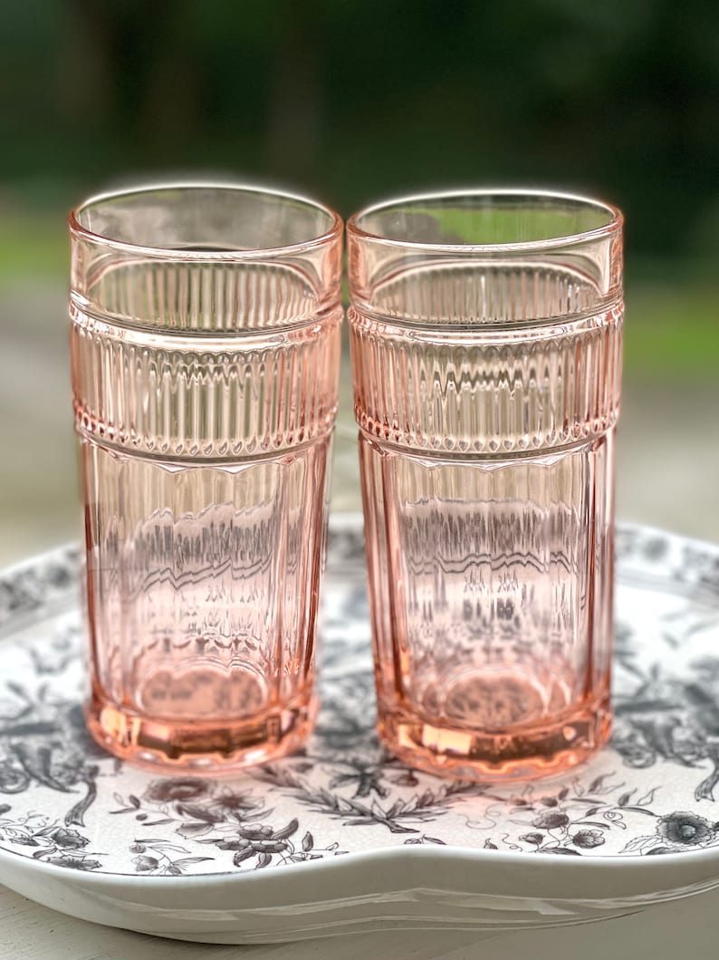 May include: Two pink glass tumblers with a ribbed design. The glasses are sitting on a white tray with a black floral pattern.
