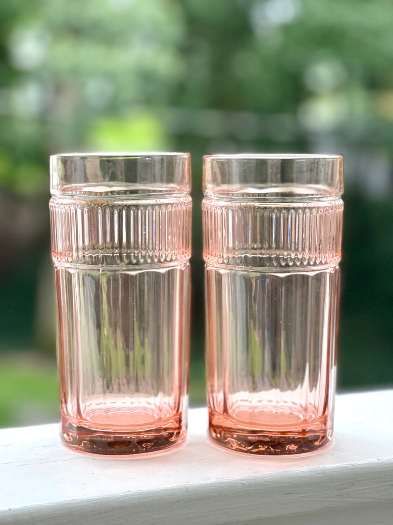 May include: Two clear pink glass tumblers with a ribbed design. The glasses are empty and sitting on a white surface.