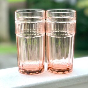 May include: Two pink glass tumblers with a ribbed design. The glasses are sitting on a white surface.