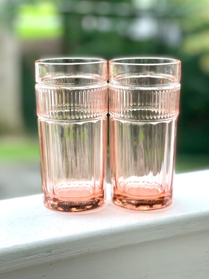May include: Two pink glass tumblers with a ribbed design. The glasses are sitting on a white surface.