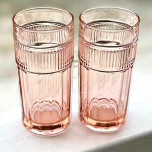 May include: Two pink glass tumblers with a ribbed design. The glasses are empty and sitting on a white surface.