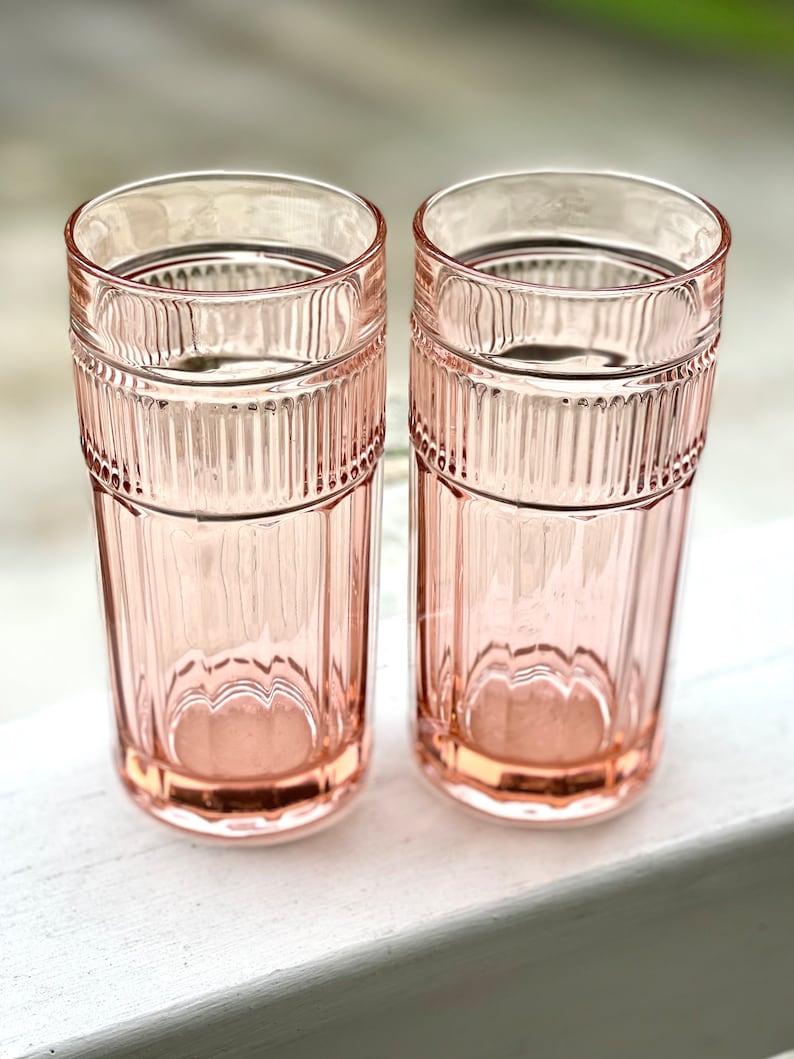 May include: Two pink glass tumblers with a ribbed design. The glasses are empty and sitting on a white surface.