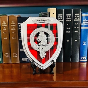 May include: A decorative shield featuring the name "MacGregor" and a clan crest, set against a red and green plaid background. The shield is displayed on a black stand in front of a row of historical books.