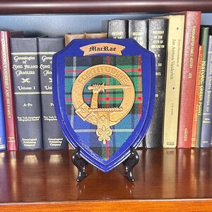 May include: A blue and green tartan shield with a gold crest and the text "MacRae" and "FORTITUDINE" on a wooden plaque.