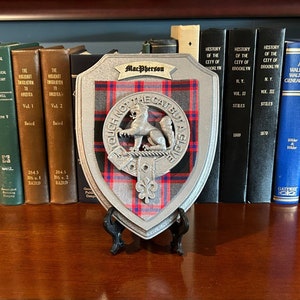 May include: A silver-colored metal shield with a red, black, and white tartan pattern. The shield has a crest with a cat and the text "Tough not the cat but a glove". The shield is on a black stand and is displayed on a shelf with books.