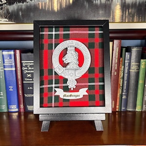 May include: A framed black and red tartan with a silver clan crest featuring a lion's head and the text "Srioghal Mo Dhream" and "MacGregor".